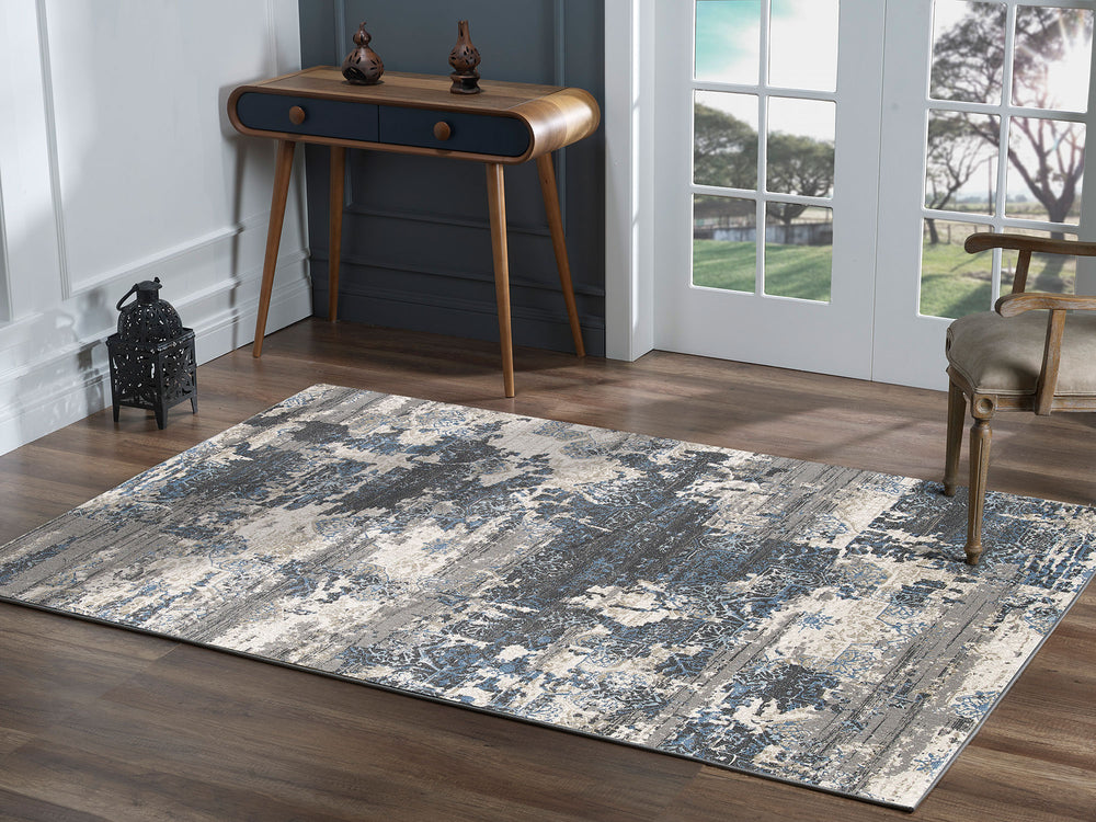A rectangular rug with a layered design featuring medium grey, deep charcoal, blue, and cream colors, and textured patterns.